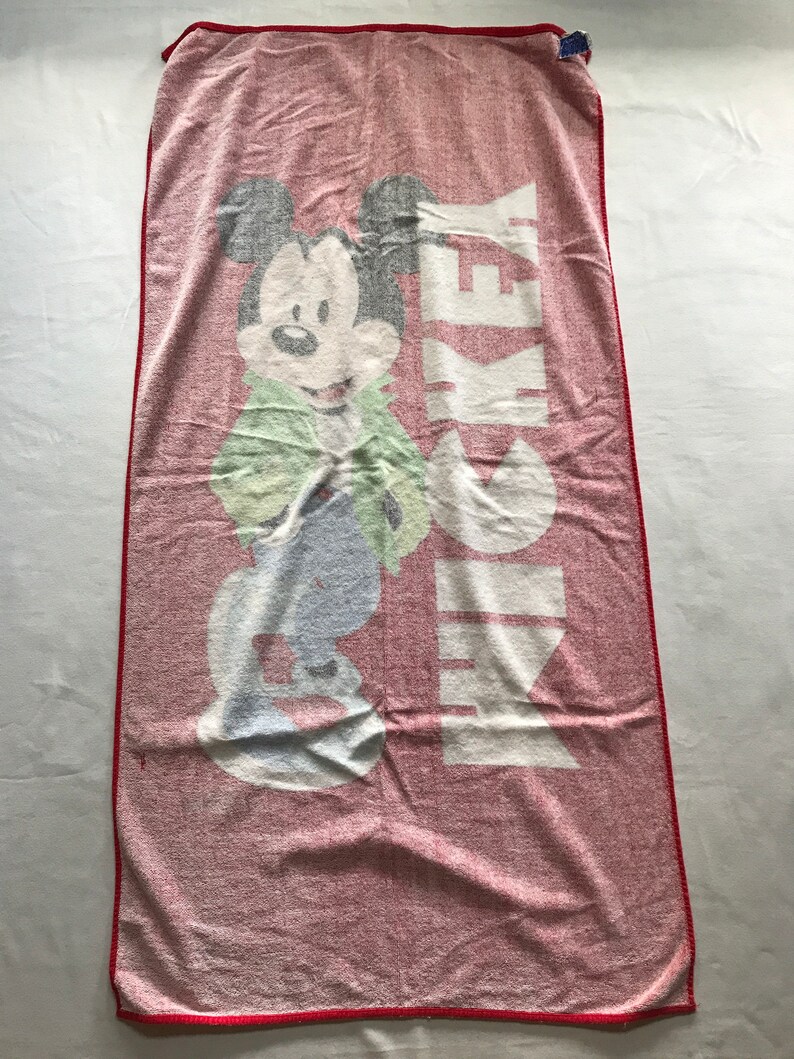 May include: A red and white Mickey Mouse beach towel. The towel features a large graphic of Mickey Mouse in a green jacket and blue pants. The word "MICKEY" is printed vertically in white. The towel has a red border.