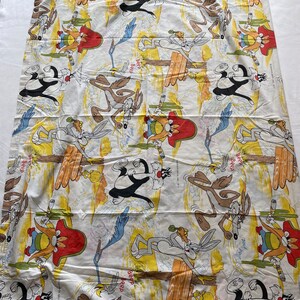 May include: A white fabric with a repeating pattern of cartoon characters from the Looney Tunes show. The characters include Wile E. Coyote, Road Runner, and Sylvester the Cat. The fabric has a yellow and brown background with the words "Beep Beep" repeated throughout.