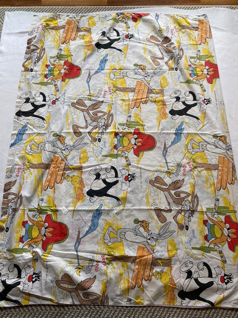 May include: A white fabric with a repeating pattern of cartoon characters from the Looney Tunes show. The characters include Wile E. Coyote, Road Runner, and Sylvester the Cat. The fabric has a yellow and brown background with the words "Beep Beep" repeated throughout.
