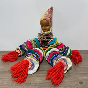 May include: A vintage handmade doll with a painted face, a floral patterned hat, and a ruffled collar. The doll's body is made of colourful fabric circles and has four red yarn tassels.
