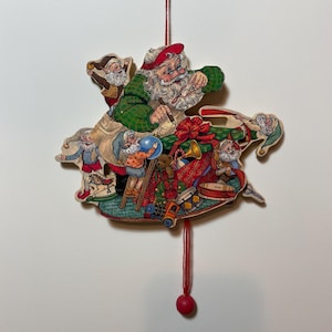 May include: A vintage Christmas ornament featuring Santa Claus and elves. Santa, in a green plaid coat and red hat, is surrounded by elves and toys. The ornament is made of wood and has a red string and a red ball at the bottom.
