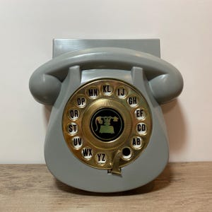 May include: A vintage gray rotary phone with a gold-colored dial. The dial features letters and numbers, with a central image of a telephone. The phone has a curved handset and a base designed for wall mounting.