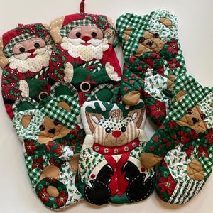 May include: A selection of Christmas-themed quilted oven mitts and pot holders. Designs include Santa Claus, reindeer, and teddy bears, featuring red, green, and white fabrics with festive patterns. Each mitt and holder has a loop for hanging.