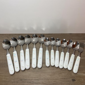 May include: A collection of stainless steel spoons and a fork with white handles. The handles are decorated with a floral pattern in blue, green, and pink. The spoons and fork are arranged on a wooden surface.