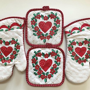 May include: A festive kitchen set comprising two oven gloves and two square pot holders. Each piece displays a white backdrop with a star pattern, a red heart, and a floral wreath. The edges are finished with red polka dots, perfect for Christmas.