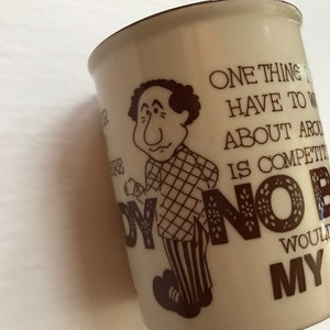 K&ouml;nnte beinhalten: Eine wei&szlig;e Keramiktasse mit einer braunen Cartoon-Illustration eines Mannes mit einem karierten Hemd und gestreiften Hosen. Die Tasse hat einen braunen Text, der lautet: "Eine Sache, &uuml;ber die ich mir hier Sorgen machen muss, ist der Wettbewerb. Niemand w&uuml;rde mit meinem..."