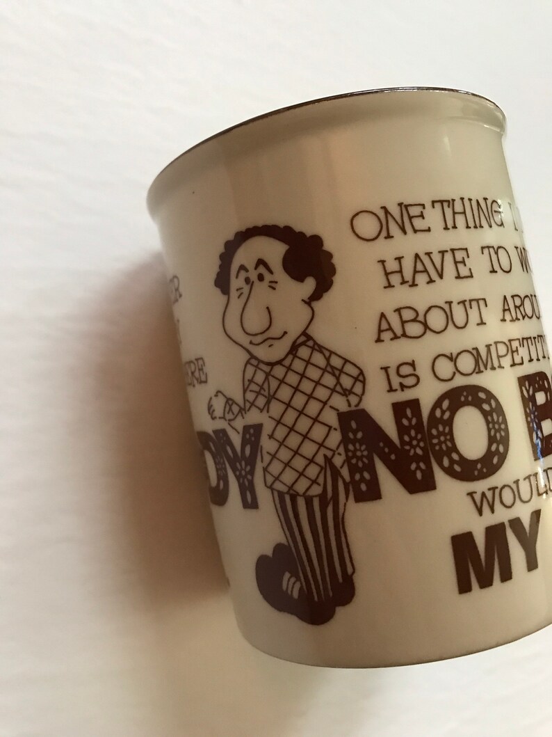K&ouml;nnte beinhalten: Eine wei&szlig;e Keramiktasse mit einer braunen Cartoon-Illustration eines Mannes mit einem karierten Hemd und gestreiften Hosen. Die Tasse hat einen braunen Text, der lautet: "Eine Sache, &uuml;ber die ich mir hier Sorgen machen muss, ist der Wettbewerb. Niemand w&uuml;rde mit meinem..."