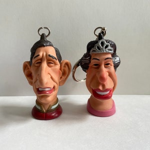 May include: A pair of keychains featuring caricature figures of a man and a woman. The man has brown hair and a large nose, while the woman has black hair and a crown. Both figures are smiling.