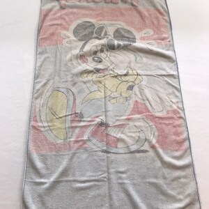 Pode incluir: Uma toalha de praia azul e branca com uma imagem de desenho animado do Mickey Mouse usando &oacute;culos de sol e uma camisa amarela. A toalha tem a palavra "Mickey" impressa em letras grandes no topo.