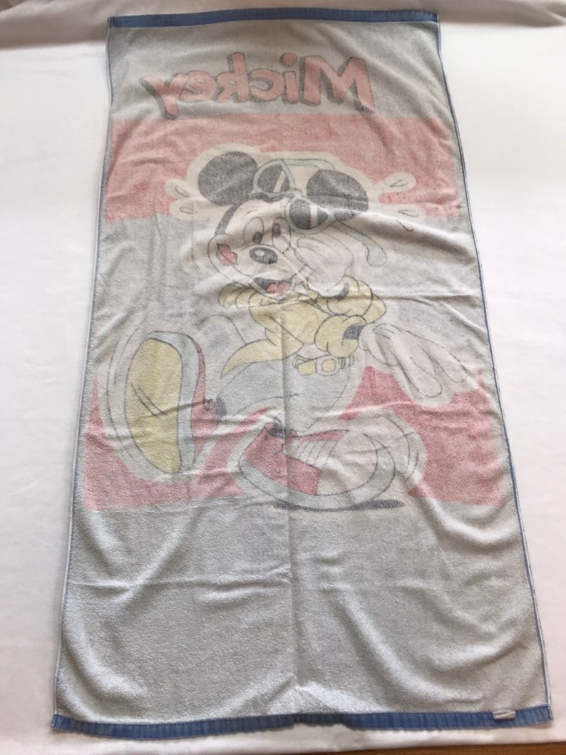 Pode incluir: Uma toalha de praia azul e branca com uma imagem de desenho animado do Mickey Mouse usando &oacute;culos de sol e uma camisa amarela. A toalha tem a palavra "Mickey" impressa em letras grandes no topo.
