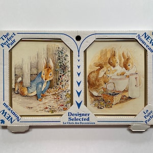 May include: A set of two framed illustrations featuring Peter Rabbit. One illustration shows Peter Rabbit wearing a blue jacket and hiding in a garden. The other illustration shows three rabbits washing dishes in a kitchen.