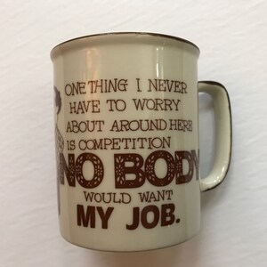K&ouml;nnte beinhalten: Eine beige Keramik-Kaffeetasse mit braunem Rand und Henkel. Die Tasse hat ein braunes Motiv einer Person und den Text "ONE THING I NEVER HAVE TO WORRY ABOUT AROUND HERE IS COMPETITION NO BODY WOULD WANT MY JOB."