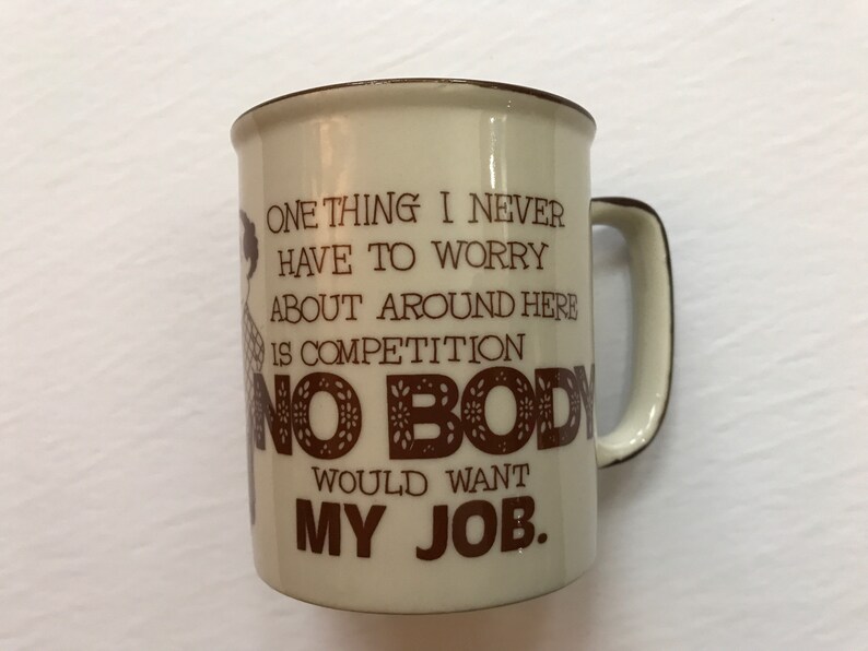 K&ouml;nnte beinhalten: Eine beige Keramik-Kaffeetasse mit braunem Rand und Henkel. Die Tasse hat ein braunes Motiv einer Person und den Text "ONE THING I NEVER HAVE TO WORRY ABOUT AROUND HERE IS COMPETITION NO BODY WOULD WANT MY JOB."
