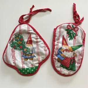 May include: Two Christmas pot holders. One is triangular, the other oval. Both have a white background with red stripes and red trim. Each features a gnome illustration. One gnome holds a Christmas tree, the other has a green hat and red polka dot apron.
