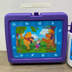 May include: Purple Winnie the Pooh lunchbox with a picture of Winnie the Pooh, Piglet, and Tigger on a blue background. The lunchbox has a white handle and a thermos logo.