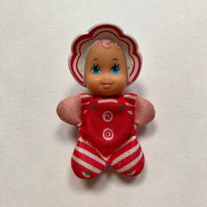 May include: A small, red and white striped doll with a pink face and blue eyes. The doll is wearing a white and red hat with a flower design.