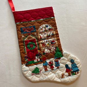 May include: Quilted Christmas stocking with a red top and a festive scene. The main design features a toy store with the word "Toys" above the door. Children are depicted playing in the snow, with a Christmas tree and a dog.