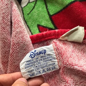 May include: A red and white Disney-themed towel with a green and red character design. The towel has a tag that reads "Disney" and "Made in Brazil". The towel is made of 100% cotton.
