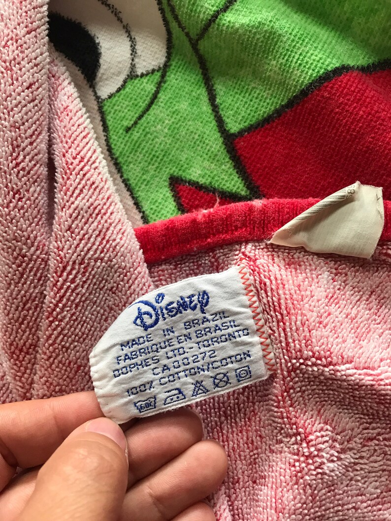 May include: A red and white Disney-themed towel with a green and red character design. The towel has a tag that reads "Disney" and "Made in Brazil". The towel is made of 100% cotton.