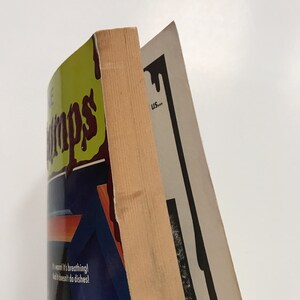Op de afbeelding: Een paperback boek met een groene kaft en de titel "The Grimps" in bruine letters. Het boek is gedeeltelijk bedekt met een blauwe en oranje sticker met de tekst "It's warm! It's breathing! And it doesn't do dishes!" Het boek ligt op een ander boek met een zwart-witte kaft.