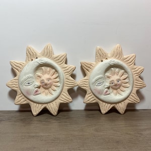 May include: Two identical circular wall plaques featuring a relief design of a crescent moon cradling a sun face. The pieces are crafted from a light beige material with subtle pink blush accents on the cheeks and lips of the celestial figures.