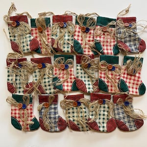 May include: A collection of miniature Christmas stockings. Each stocking features a red, green, and white plaid pattern, with a green cuff and red heel. They are adorned with twine bows and wooden buttons, and are attached to twine with small wooden clips.