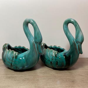 May include: Two teal blue ceramic swan planters with a glazed finish. The swans have a detailed design and are perfect for displaying small plants or flowers.