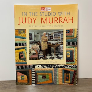 May include: A book titled "In The Studio With Judy Murrah" with the subtitle "12 Playful Quilted Projects." The cover shows a woman in a studio, surrounded by colourful quilt blocks. The book is a guide to quilting.