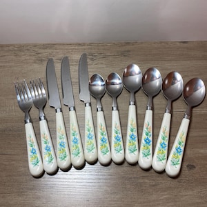 May include: A collection of vintage silverware, including forks, knives, and spoons. The handles are white with a floral pattern in blue, yellow, and green. The stainless steel utensils are arranged on a wooden surface.