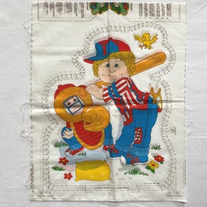 May include: A white fabric pattern with a boy wearing a red and white striped shirt and a baseball cap holding a bat. The boy is standing next to a yellow and red catcher's mitt. The pattern includes instructions for making a stuffed toy.