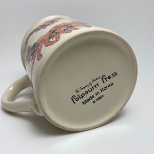 May include: Cream-colored ceramic mug with a floral design in pink, orange, and green. The mug has a curved handle and the text "Potpourri Press Made in Korea &copy;1989" printed on the bottom. The mug is angled, showing the bottom and side.
