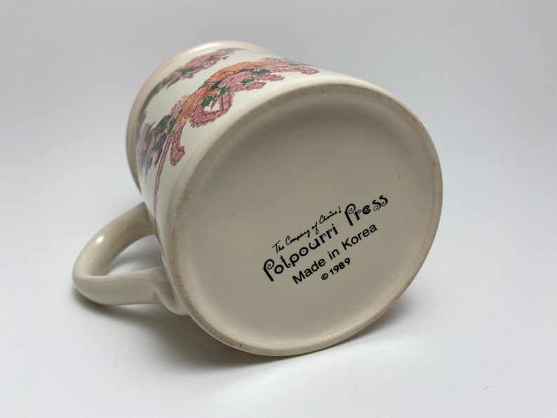 May include: Cream-colored ceramic mug with a floral design in pink, orange, and green. The mug has a curved handle and the text "Potpourri Press Made in Korea &copy;1989" printed on the bottom. The mug is angled, showing the bottom and side.