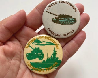 May include: Two vintage Canadian Forces buttons. One button is white with a green tank and the text "Forces Canadiennes" and "Canadian Forces". The other button is beige with a green tank, a green jet, and a green ship with the text "Forces Canadiennes" and "Canadian Forces".