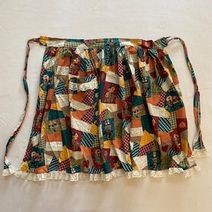 May include: A colorful patchwork apron with a variety of patterns and colors, including red, green, yellow, and orange. The apron has a white lace trim along the bottom and long ties for securing around the waist.