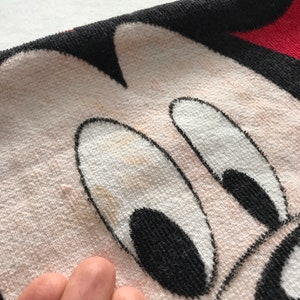 May include: Close-up of a white and black Mickey Mouse face on a towel. The towel has a red border and black outline. The face has black eyes and a black nose. The towel is made of a soft, absorbent material.