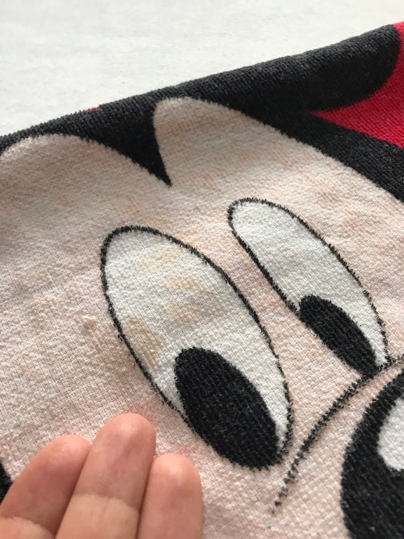 May include: Close-up of a white and black Mickey Mouse face on a towel. The towel has a red border and black outline. The face has black eyes and a black nose. The towel is made of a soft, absorbent material.