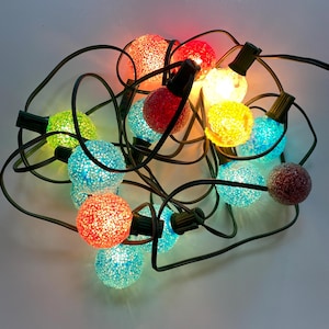 May include: A string of colorful Christmas lights with textured, spherical bulbs. The lights are in various colors, including red, blue, yellow, and green, and are connected by a black wire. The bulbs have a glittery appearance.