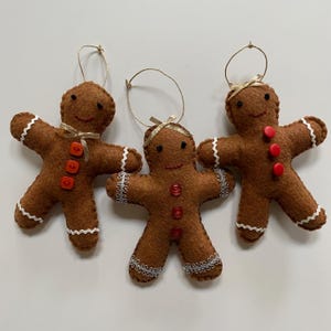 May include: Three felt gingerbread people ornaments with white trim and red buttons. Each ornament has a gold ribbon tied around its neck.