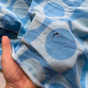 May include: A close-up of a light blue fabric with a white and light blue geometric pattern. The fabric has a small hole in the centre of one of the blue circles.