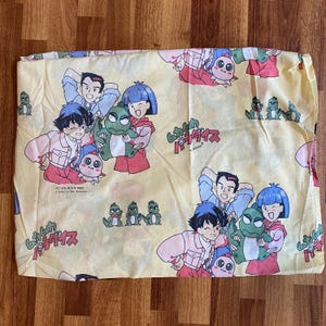 May include: A light yellow fabric sheet featuring a repeating pattern of anime characters, including a green creature, a person with dark hair in a pink jacket, and a person with blue hair in a red jacket, with Japanese text and the year 1993 printed.