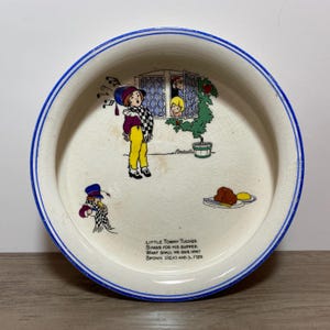 Peut inclure: Une assiette ronde en céramique avec une base crème et un bord bleu. Le centre présente une illustration colorée du personnage Little Tommy Tucker, une fenêtre, une plante et de la nourriture. Le texte indique : Little Tommy Tucker Sings for his supper. What shall we give him? Brown bread and butter.