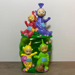May include: A green ceramic jar featuring the Teletubbies characters. The lid has a red and purple Teletubby sitting on a green base with flowers. The jar has yellow and green Teletubbies with a pink watering can and a brown rabbit.