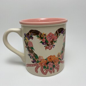 May include: Cream-colored ceramic mug with a heart-shaped floral design. The heart is composed of pink and orange roses, purple flowers, and brown ribbons. The rim and interior of the mug are pink, and it has a curved handle.