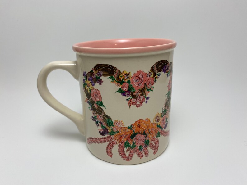 May include: Cream-colored ceramic mug with a heart-shaped floral design. The heart is composed of pink and orange roses, purple flowers, and brown ribbons. The rim and interior of the mug are pink, and it has a curved handle.