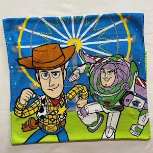 Puede incluir: Una manta de franela azul y verde con una imagen colorida de Woody y Buzz Lightyear de la franquicia de pel&iacute;culas Toy Story. Los personajes est&aacute;n de pie frente a una noria.