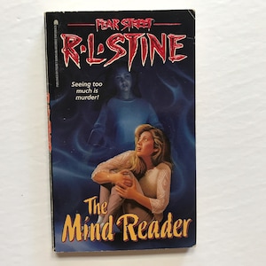 May include: A paperback book cover with a black background and a white title that reads "The Mind Reader". The author's name, R.L. Stine, is printed in white above the title. The cover features a young woman with long blonde hair looking up in fear. A ghostly figure of a young girl with long dark hair is behind her.