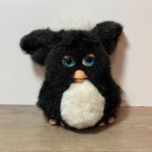 May include: A vintage black Furby plush toy with blue eyes, a white belly, and a tuft of white fur on top. The Furby has a tan beak and small feet. The plush toy is sitting on a wooden surface.