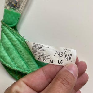 May include: A close-up of a green toy with a tag. The tag has the text "2436HR" written on it. The toy is made of green fabric and has a clear plastic piece with glitter.