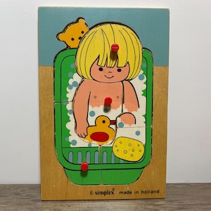 May include: A vintage wooden puzzle depicting a child in a bathtub with a rubber duck and a sponge. The puzzle has a light blue background and a green bathtub. The child has yellow hair and is surrounded by bubbles. The puzzle is marked "simplex made in holland."