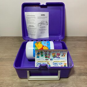 May include: Purple Winnie the Pooh lunchbox with a white thermos inside. The lunchbox has a white handle and a white latch. The thermos has a Winnie the Pooh design on it. There are coupons for Johnson & Johnson products inside the lunchbox.
